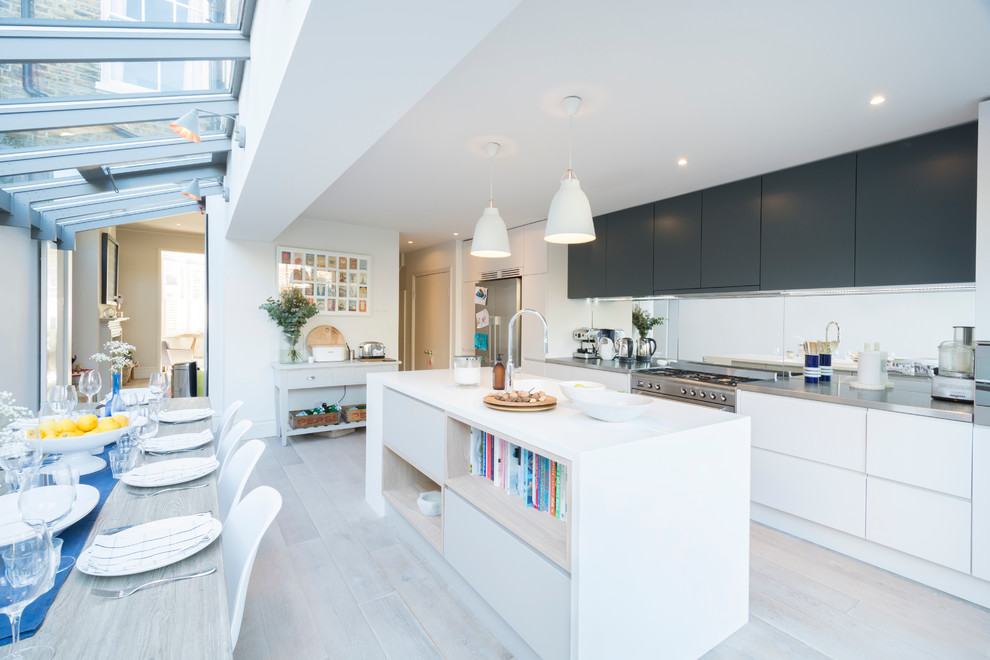 Photo of a medium sized scandinavian kitchen/diner in London with a double-bowl sink, flat-panel cabinets, blue cabinets, stainless steel worktops, metallic splashback, mirror splashback, stainless steel appliances, light hardwood flooring and an island.