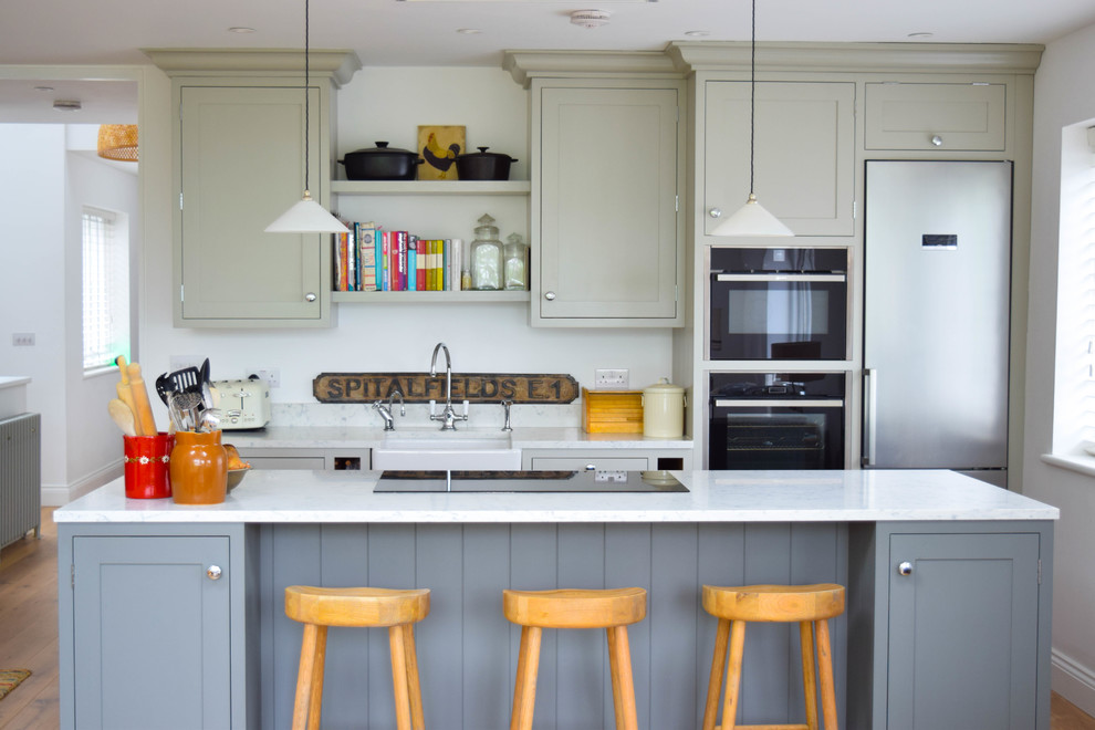 This is an example of a large rural single-wall kitchen in London with a belfast sink, shaker cabinets, marble worktops, white splashback, marble splashback, stainless steel appliances, light hardwood flooring, an island, white worktops and grey cabinets.