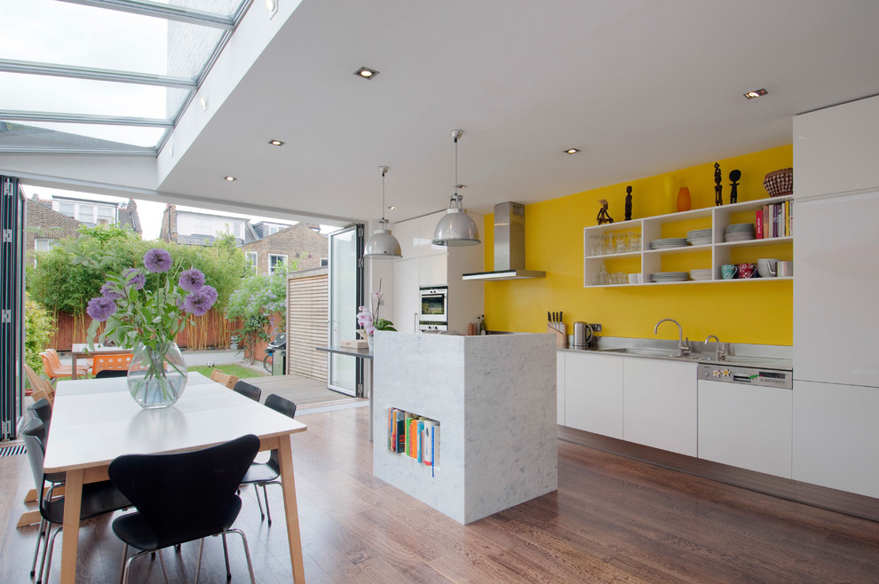 Moderne Wohnküche mit integriertem Waschbecken, weißen Schränken, Edelstahl-Arbeitsplatte, Küchenrückwand in Gelb, Elektrogeräten mit Frontblende, dunklem Holzboden, Kücheninsel, braunem Boden und offenen Schränken in London