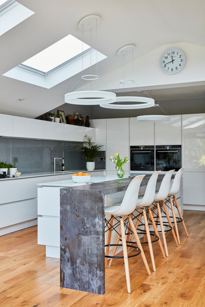 Inspiration for a large contemporary l-shaped kitchen/diner in London with flat-panel cabinets, white cabinets, granite worktops, grey splashback, an island, brown floors, white worktops, black appliances, medium hardwood flooring and a vaulted ceiling.