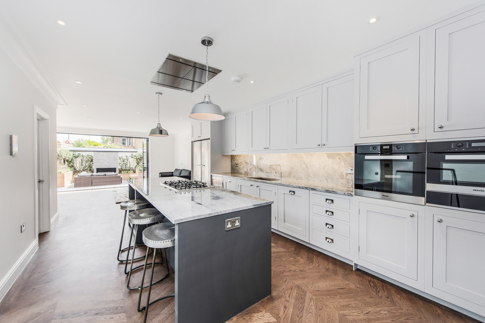 Inspiration for a large traditional single-wall kitchen/diner in London with recessed-panel cabinets, white cabinets, granite worktops, stone slab splashback, medium hardwood flooring, an island, an integrated sink and stainless steel appliances.