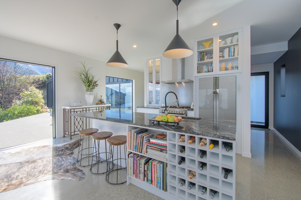 This is an example of a large contemporary galley kitchen in Other with glass-front cabinets, white cabinets, white splashback, glass sheet splashback, stainless steel appliances, concrete flooring, an island, grey worktops, a submerged sink and grey floors.