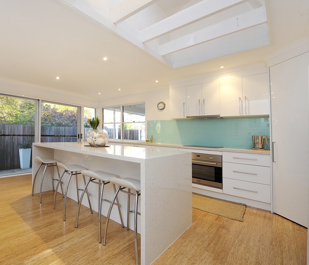 Moderne Küche mit flächenbündigen Schrankfronten, weißen Schränken, Küchenrückwand in Blau, Küchengeräten aus Edelstahl, braunem Holzboden und Kücheninsel in Brisbane