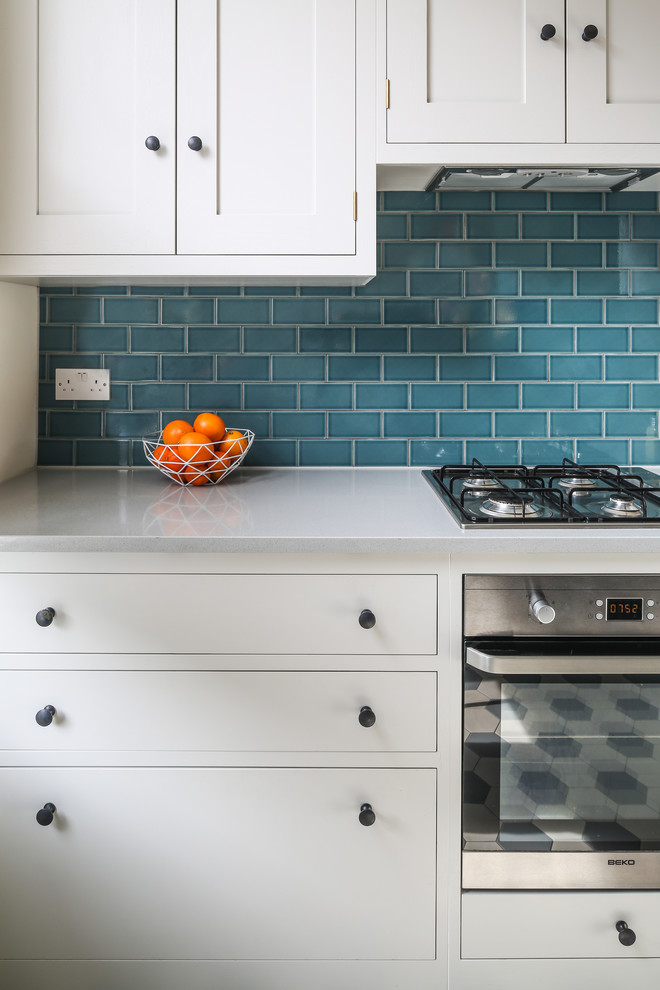 Example of a small trendy l-shaped ceramic tile and multicolored floor open concept kitchen design in London with a farmhouse sink, shaker cabinets, white cabinets, quartz countertops, blue backsplash, subway tile backsplash, stainless steel appliances and a peninsula