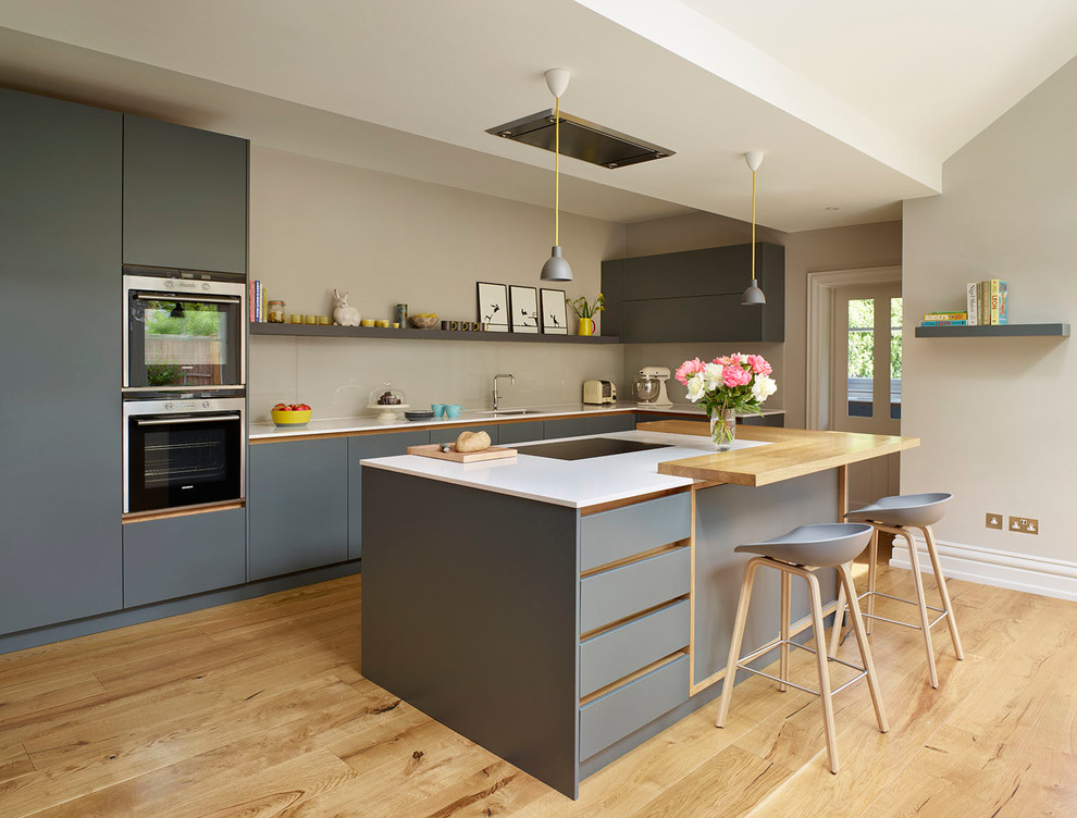 Photo of a large scandi l-shaped kitchen in London with flat-panel cabinets, grey cabinets, composite countertops, beige splashback, light hardwood flooring, an island, beige floors, white worktops, a submerged sink, glass sheet splashback and stainless steel appliances.