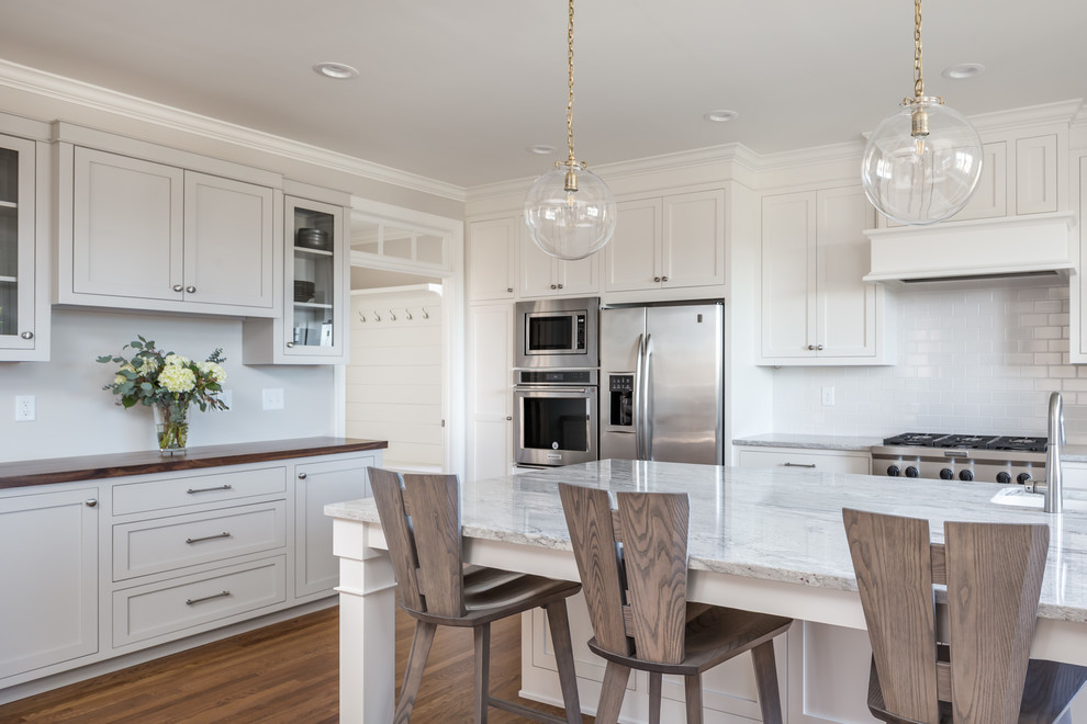 Exemple d'une grande cuisine américaine chic en U avec un placard à porte shaker, des portes de placard blanches, une crédence blanche, un électroménager en acier inoxydable, un sol en bois brun, îlot, un évier de ferme, un sol marron, un plan de travail en quartz, une crédence en carrelage métro et un plan de travail blanc.