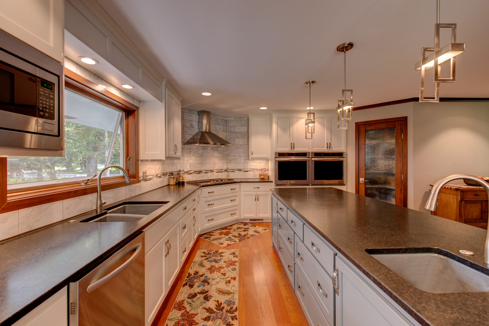 Example of a large transitional l-shaped medium tone wood floor and brown floor kitchen pantry design in Minneapolis with an undermount sink, recessed-panel cabinets, white cabinets, granite countertops, white backsplash, stone tile backsplash, stainless steel appliances and an island