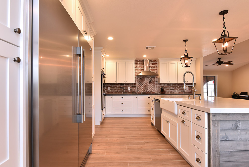 Bild på ett stort lantligt kök, med en rustik diskho, skåp i shakerstil, vita skåp, bänkskiva i kvarts, rött stänkskydd, stänkskydd i tegel, rostfria vitvaror, ljust trägolv, en köksö och brunt golv
