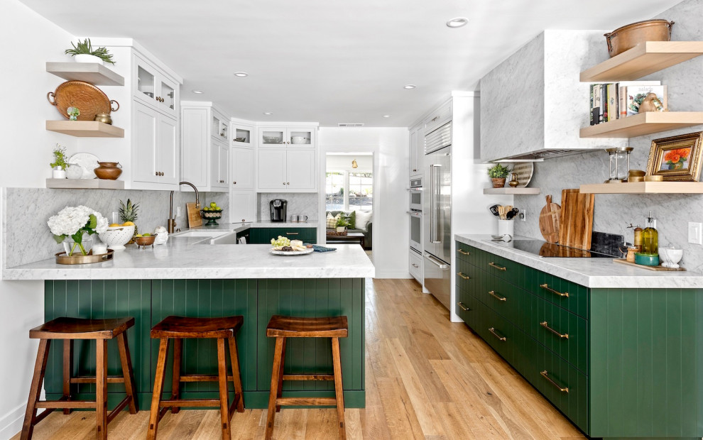 This is an example of a classic u-shaped kitchen in San Diego with a belfast sink, flat-panel cabinets, green cabinets, white splashback, stone slab splashback, stainless steel appliances, medium hardwood flooring, a breakfast bar, brown floors and white worktops.