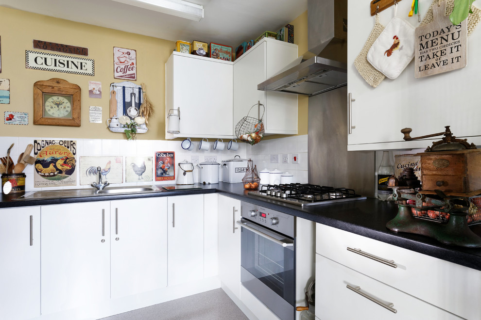 This is an example of a small shabby-chic style l-shaped enclosed kitchen in London with a built-in sink, flat-panel cabinets, white cabinets, laminate countertops, metallic splashback, stainless steel appliances, no island and grey floors.