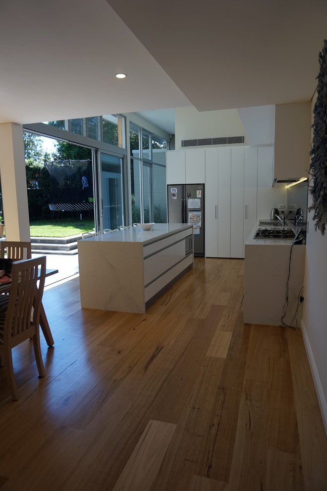 Inspiration for a medium sized modern l-shaped kitchen pantry in Sydney with a built-in sink, white cabinets, engineered stone countertops, glass sheet splashback, stainless steel appliances, light hardwood flooring and an island.