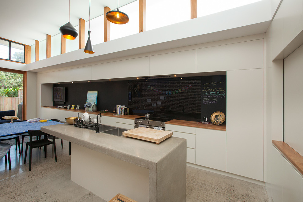 На фото: параллельная кухня-гостиная среднего размера в современном стиле с монолитной мойкой, белыми фасадами, столешницей из бетона, черным фартуком, фартуком из стекла, бетонным полом, островом и техникой под мебельный фасад