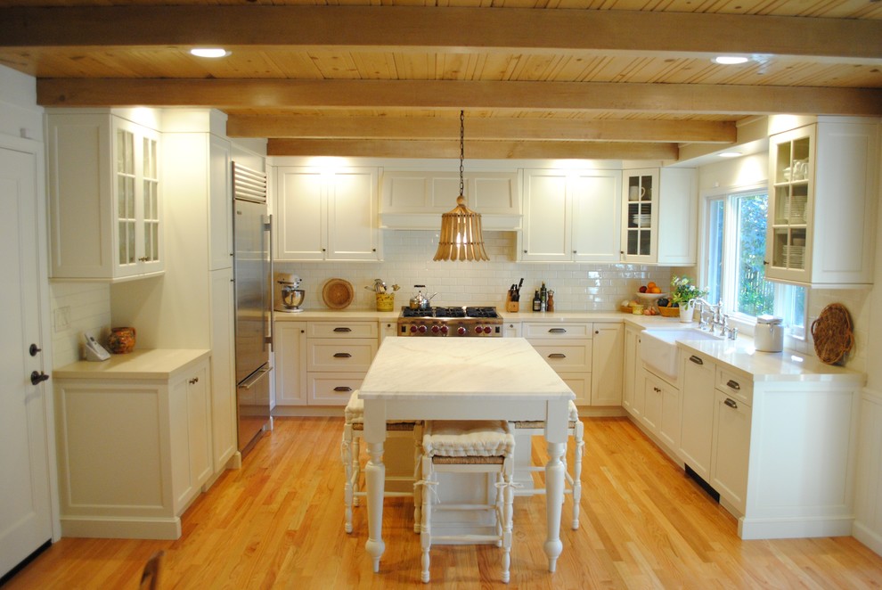 Photo of a large rural u-shaped kitchen/diner in San Francisco with a belfast sink, shaker cabinets, white cabinets, marble worktops, white splashback, metro tiled splashback, stainless steel appliances, light hardwood flooring and an island.