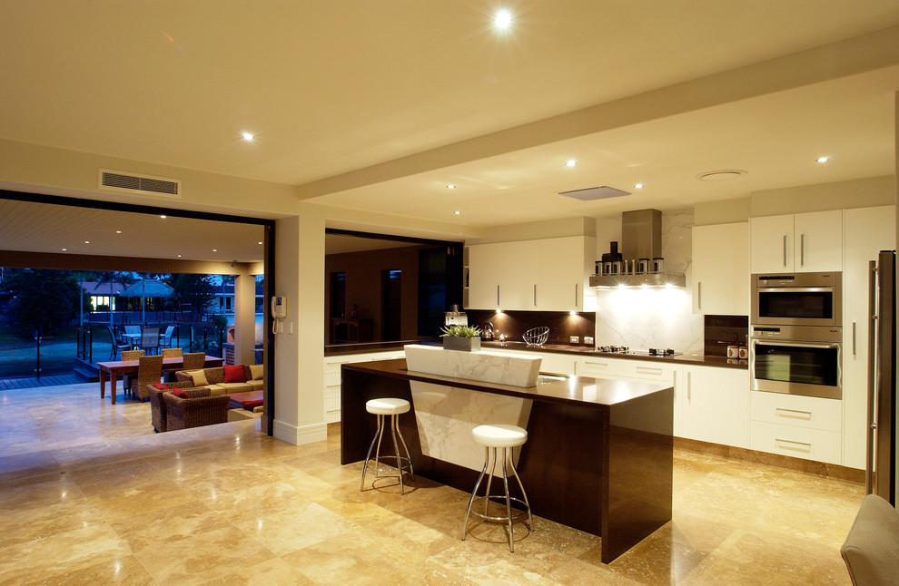 This is an example of a large contemporary l-shaped kitchen/diner in Brisbane with a submerged sink, flat-panel cabinets, white cabinets, white splashback, marble splashback, stainless steel appliances, travertine flooring, an island and beige floors.