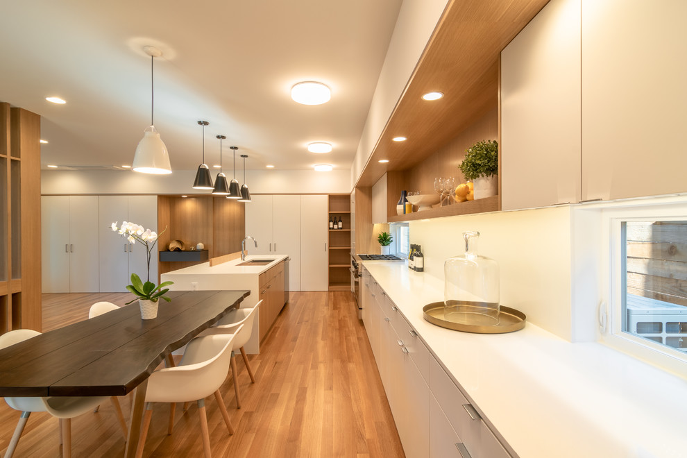 Medium sized modern single-wall open plan kitchen in Seattle with a submerged sink, flat-panel cabinets, white cabinets, composite countertops, white splashback, stone slab splashback, stainless steel appliances, medium hardwood flooring, an island, brown floors and white worktops.