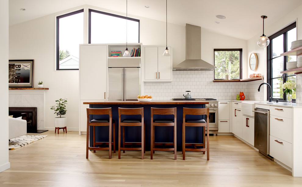 Modern inredning av ett vit vitt kök, med en rustik diskho, släta luckor, blå skåp, träbänkskiva, vitt stänkskydd, rostfria vitvaror, ljust trägolv, en köksö och beiget golv