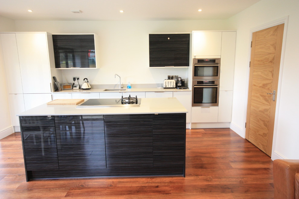 Photo of a large contemporary single-wall open plan kitchen in Kent with an integrated sink, flat-panel cabinets, white cabinets, quartz worktops, stainless steel appliances, medium hardwood flooring and an island.