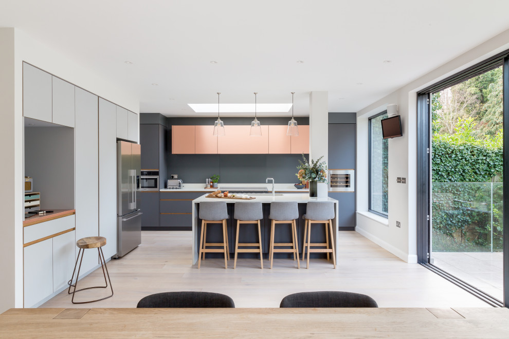 Example of a large trendy single-wall medium tone wood floor and beige floor open concept kitchen design in London with a double-bowl sink, flat-panel cabinets, gray cabinets, solid surface countertops, gray backsplash, stainless steel appliances, an island and white countertops