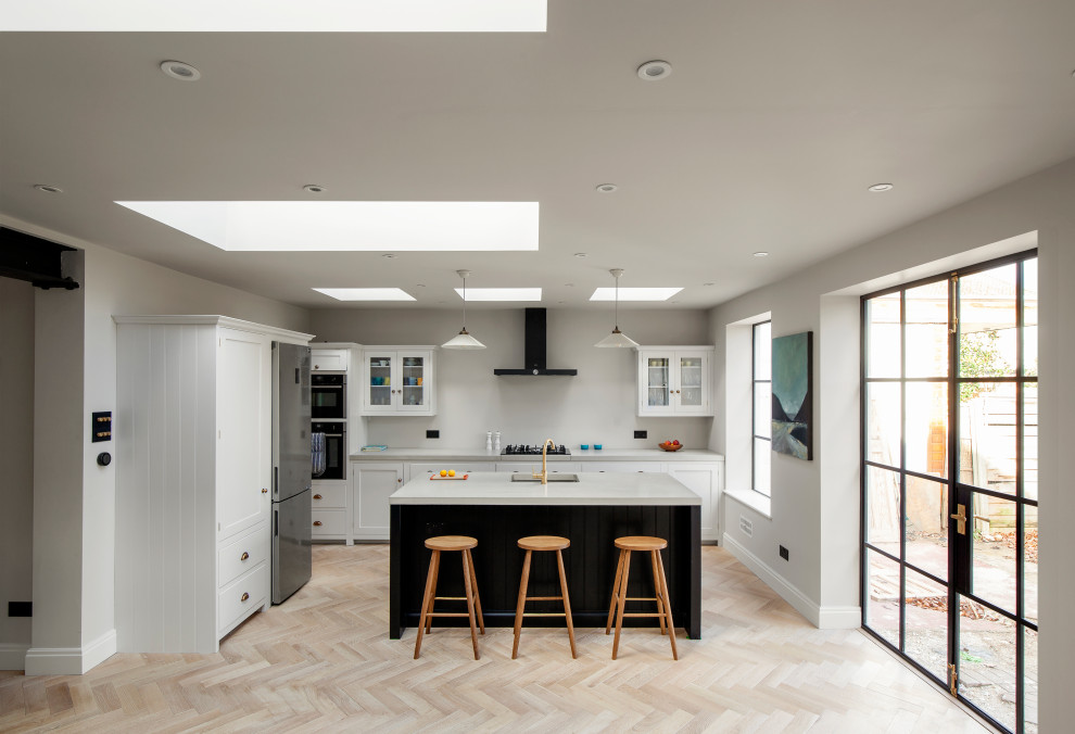 This is an example of a medium sized classic l-shaped open plan kitchen in London with shaker cabinets, concrete worktops, light hardwood flooring, an island, grey worktops, white cabinets, stainless steel appliances and beige floors.