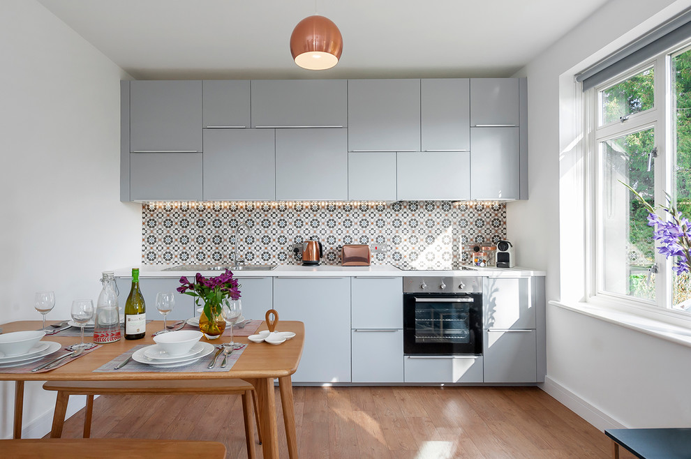 Small trendy single-wall light wood floor and brown floor eat-in kitchen photo in Wiltshire with flat-panel cabinets, blue cabinets, stainless steel appliances, a drop-in sink and multicolored backsplash