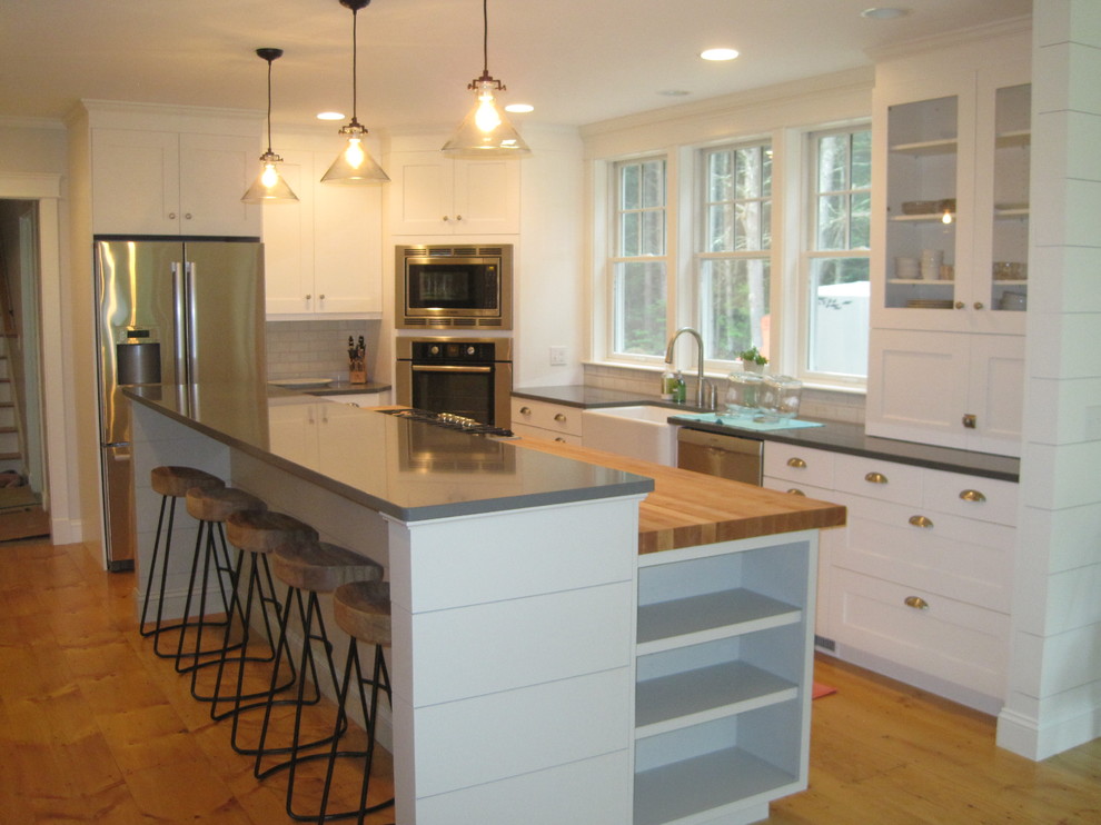 This is an example of a large classic u-shaped kitchen in Portland Maine with a belfast sink, shaker cabinets, white cabinets, white splashback, metro tiled splashback, stainless steel appliances, medium hardwood flooring, an island, brown floors and black worktops.
