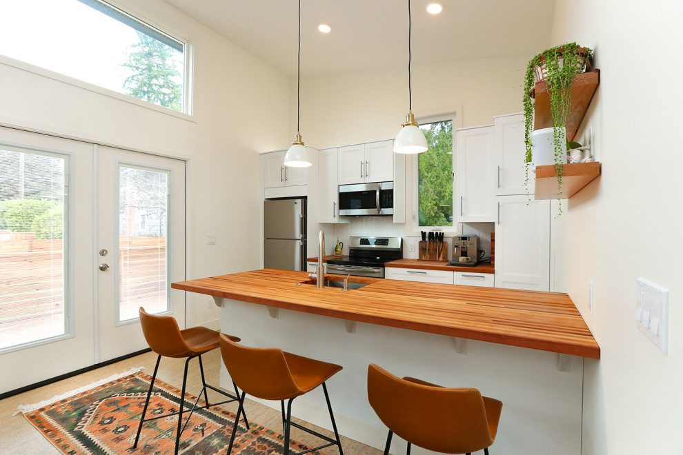 Example of a small danish galley cork floor open concept kitchen design in Seattle with an undermount sink, shaker cabinets, white cabinets, wood countertops, white backsplash, subway tile backsplash, stainless steel appliances and a peninsula