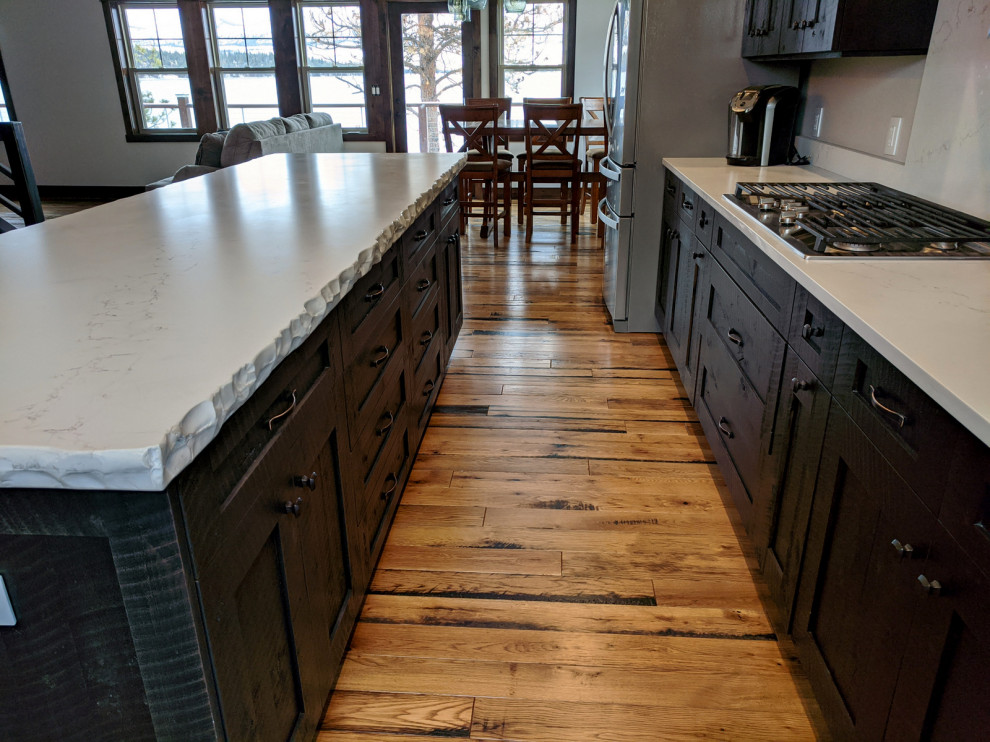 Large mountain style l-shaped medium tone wood floor and brown floor open concept kitchen photo in Other with a farmhouse sink, recessed-panel cabinets, dark wood cabinets, quartzite countertops, white backsplash, travertine backsplash, stainless steel appliances and white countertops
