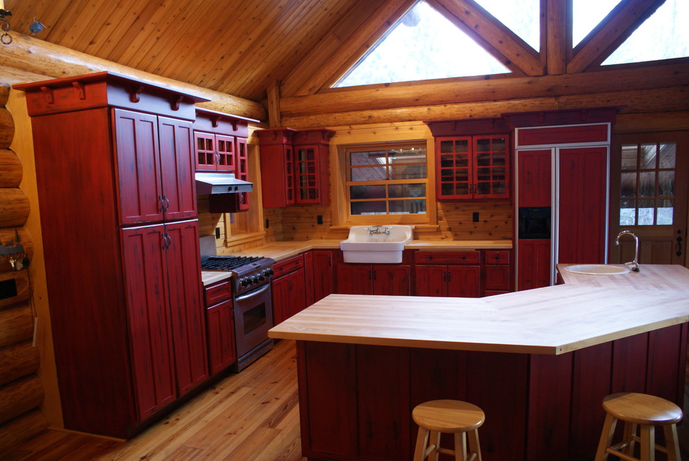 他の地域にある広いラスティックスタイルのおしゃれなキッチン (落し込みパネル扉のキャビネット、赤いキャビネット、エプロンフロントシンク、木材カウンター、茶色いキッチンパネル、木材のキッチンパネル、パネルと同色の調理設備、無垢フローリング、茶色い床) の写真