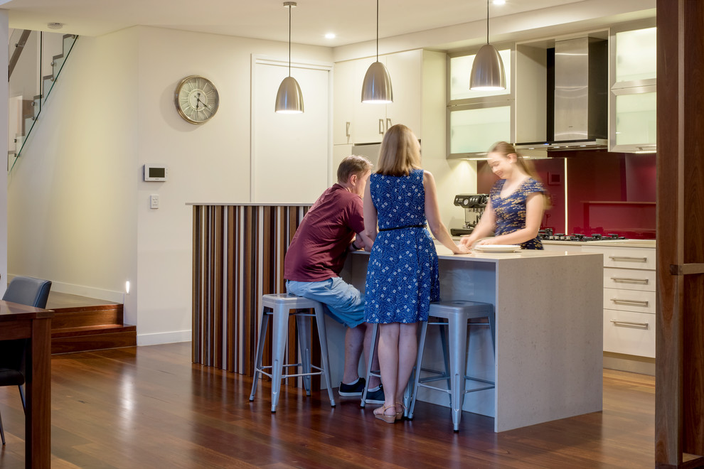 Large contemporary galley open plan kitchen in Brisbane with a double-bowl sink, red splashback, glass sheet splashback, stainless steel appliances, medium hardwood flooring, an island, flat-panel cabinets, white cabinets, quartz worktops and brown floors.