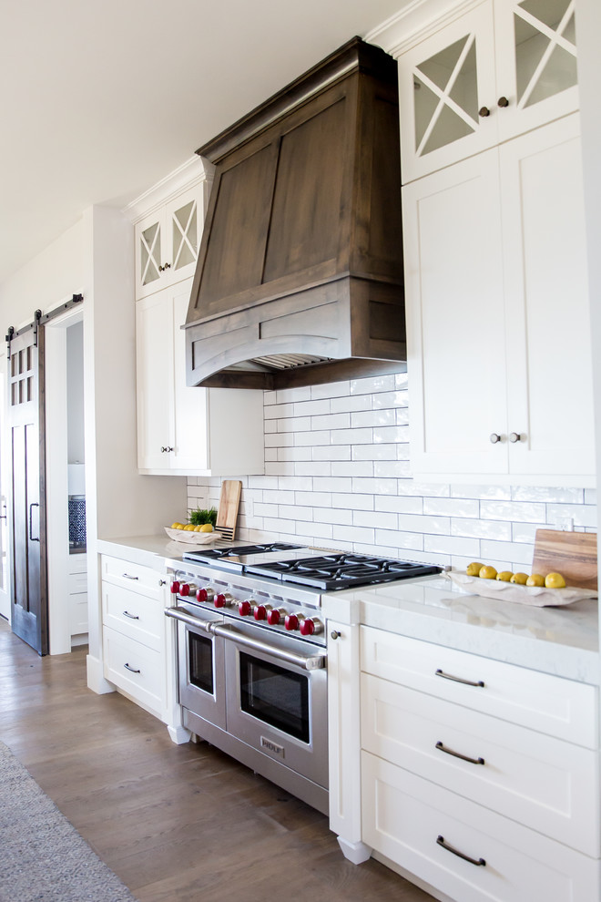 This is an example of a large farmhouse single-wall open plan kitchen in Salt Lake City with a belfast sink, shaker cabinets, white cabinets, soapstone worktops, white splashback, metro tiled splashback, stainless steel appliances, medium hardwood flooring and an island.