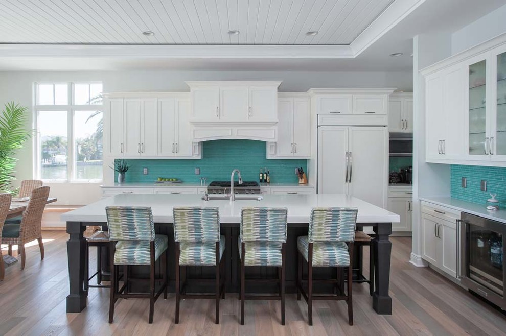 Large nautical l-shaped open plan kitchen in Other with a single-bowl sink, shaker cabinets, white cabinets, composite countertops, green splashback, matchstick tiled splashback, integrated appliances, medium hardwood flooring, an island, brown floors and white worktops.