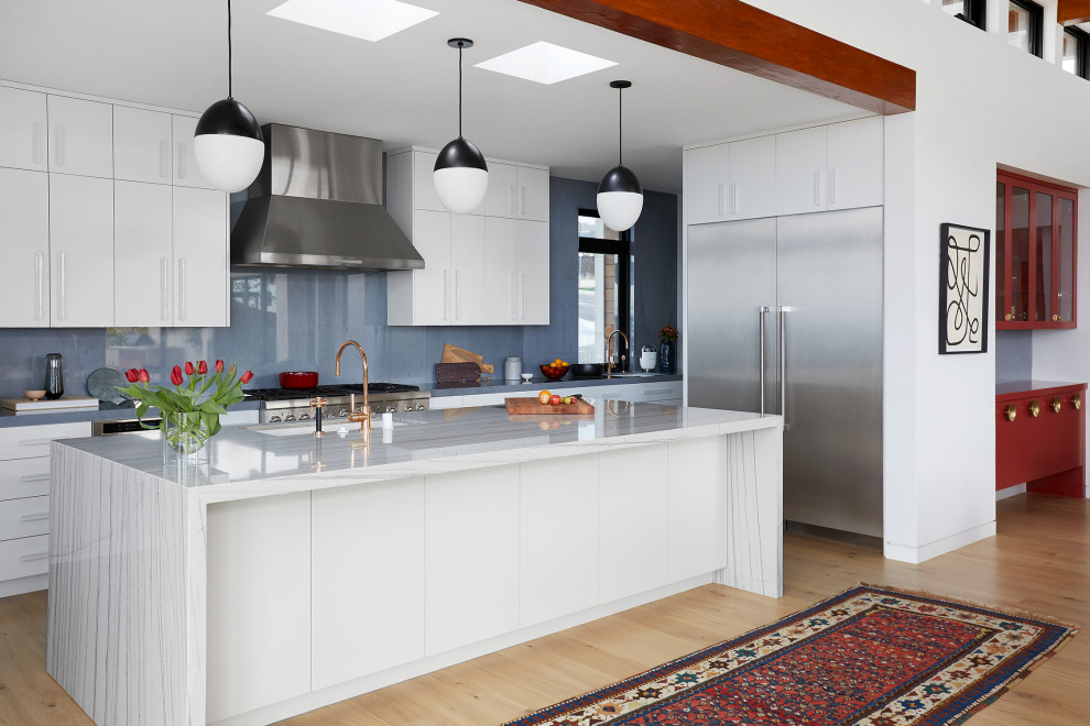 Foto på ett funkis vit kök, med släta luckor, vita skåp, marmorbänkskiva, blått stänkskydd, stänkskydd i marmor, rostfria vitvaror, ljust trägolv, en köksö och beiget golv