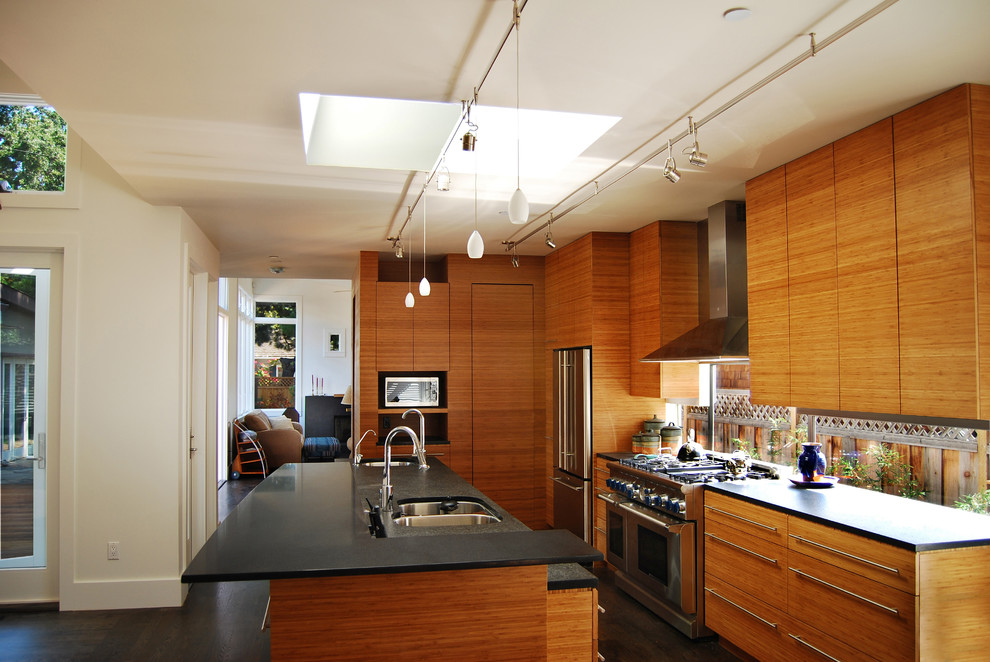 Inredning av ett modernt stort kök, med en trippel diskho, släta luckor, skåp i mellenmörkt trä, träbänkskiva, glaspanel som stänkskydd, rostfria vitvaror, mörkt trägolv, en köksö och brunt golv
