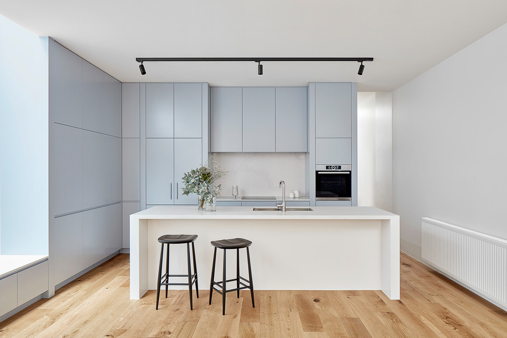 Ejemplo de cocina moderna, pequeña, con fregadero de doble seno, armarios con paneles lisos, puertas de armario azules, salpicadero blanco, suelo de madera clara, una isla, encimeras blancas, electrodomésticos con paneles y suelo beige