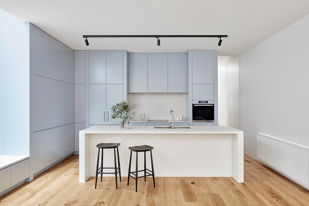 Inspiration for a small modern galley kitchen in Melbourne with a double-bowl sink, flat-panel cabinets, blue cabinets, white splashback, light hardwood flooring, an island, white worktops, integrated appliances and beige floors.