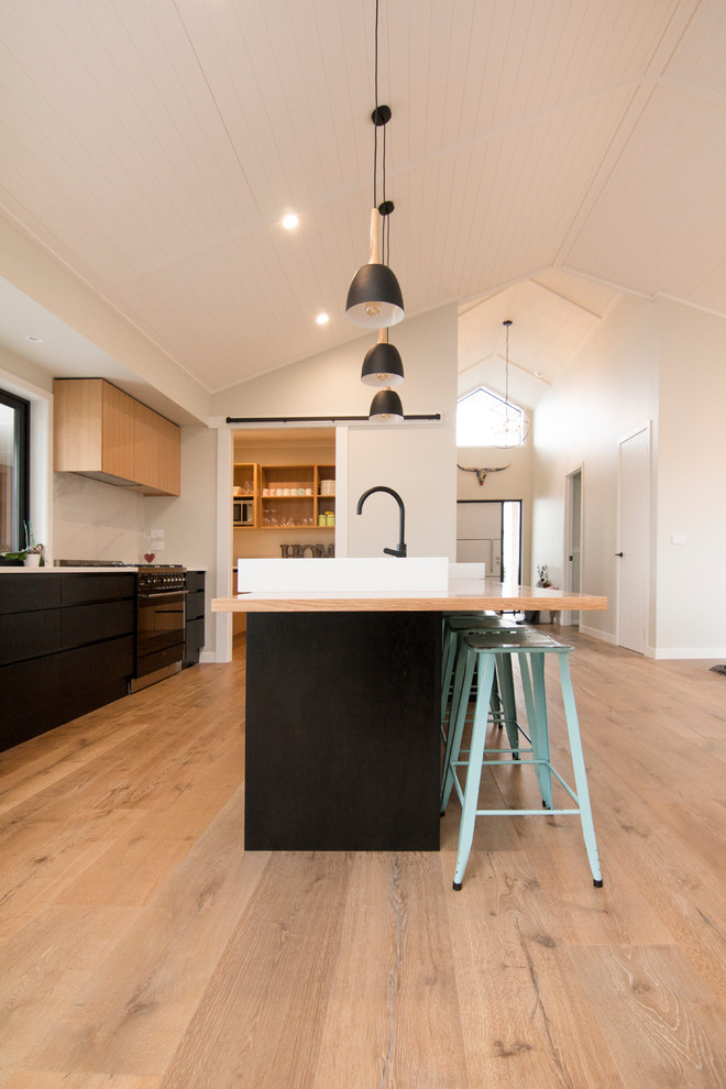 Example of a mid-sized mountain style galley medium tone wood floor and beige floor eat-in kitchen design in Auckland with a farmhouse sink, flat-panel cabinets, light wood cabinets, wood countertops, white backsplash, marble backsplash, black appliances and an island
