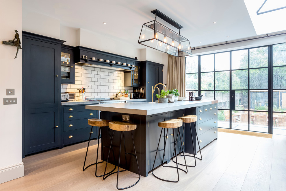 This is an example of a contemporary galley kitchen in London with shaker cabinets, black cabinets, white splashback, metro tiled splashback, integrated appliances, light hardwood flooring, an island and beige floors.