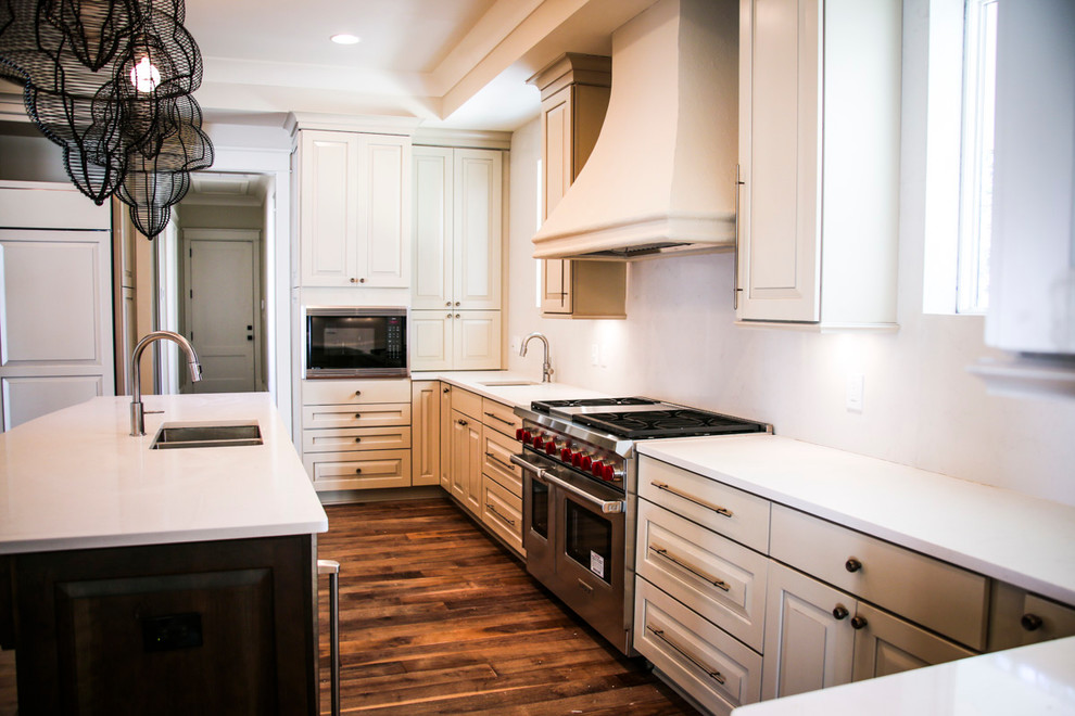 Example of a mid-sized classic l-shaped medium tone wood floor enclosed kitchen design in Miami with raised-panel cabinets, white cabinets, solid surface countertops, stainless steel appliances, an island, white backsplash and a double-bowl sink