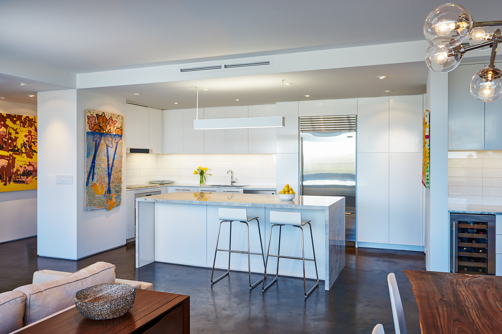 Modern inredning av ett kök, med släta luckor, vita skåp, vitt stänkskydd, rostfria vitvaror och en köksö