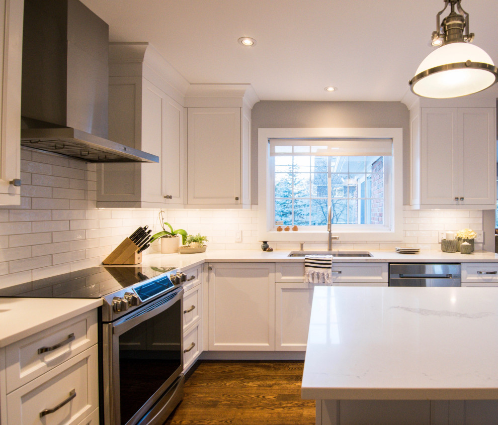 Lantlig inredning av ett stort vit vitt kök, med en undermonterad diskho, skåp i shakerstil, vita skåp, bänkskiva i kvarts, vitt stänkskydd, stänkskydd i keramik, rostfria vitvaror, mellanmörkt trägolv, en köksö och brunt golv