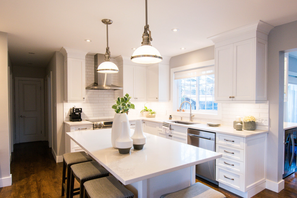 This is an example of a large farmhouse u-shaped kitchen/diner in Montreal with a submerged sink, shaker cabinets, white cabinets, engineered stone countertops, white splashback, ceramic splashback, stainless steel appliances, medium hardwood flooring, an island, brown floors and white worktops.