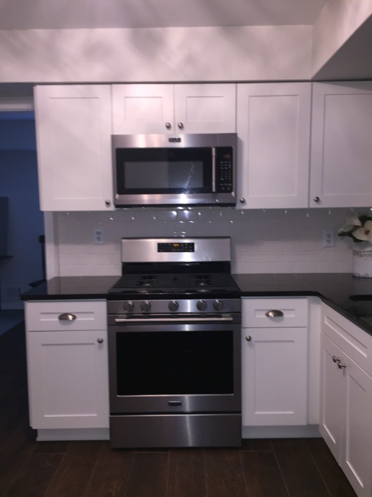 ニューヨークにある低価格の中くらいなトラディショナルスタイルのおしゃれなL型キッチン (アンダーカウンターシンク、シェーカースタイル扉のキャビネット、白いキャビネット、クオーツストーンカウンター、白いキッチンパネル、磁器タイルのキッチンパネル、シルバーの調理設備、磁器タイルの床、アイランドなし、茶色い床、黒いキッチンカウンター) の写真