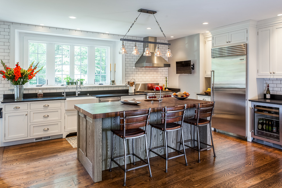This is an example of a classic u-shaped kitchen in Philadelphia with a belfast sink, shaker cabinets, white cabinets, white splashback, metro tiled splashback, stainless steel appliances, medium hardwood flooring, an island and brown floors.