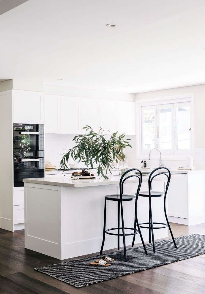 Design ideas for a medium sized classic l-shaped open plan kitchen in Sydney with shaker cabinets, white cabinets, white splashback, medium hardwood flooring, an island, brown floors and white worktops.