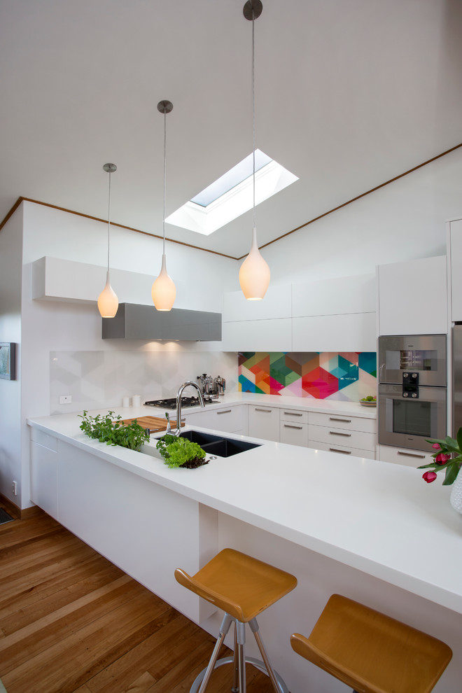 Exempel på ett modernt kök, med en dubbel diskho, släta luckor, vita skåp, stänkskydd med metallisk yta, rostfria vitvaror, mellanmörkt trägolv och en halv köksö