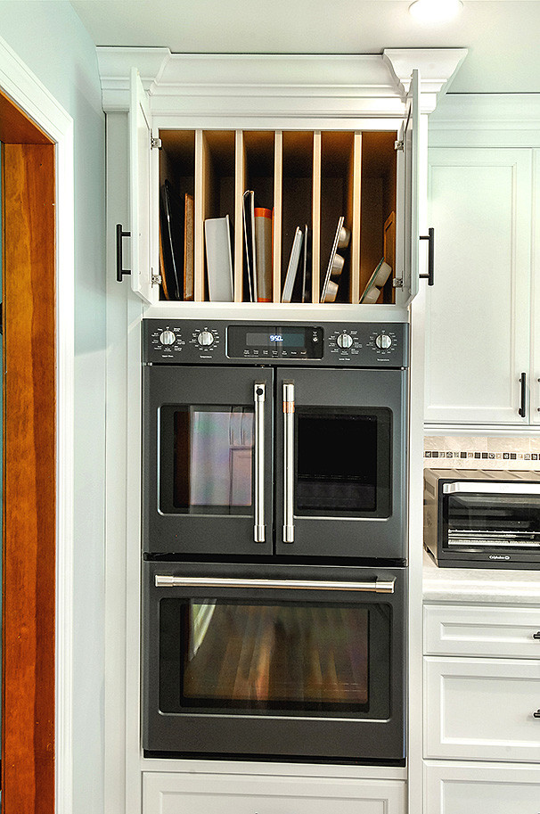 This is an example of a traditional kitchen/diner in Philadelphia with a belfast sink, recessed-panel cabinets, white cabinets, engineered stone countertops, grey splashback, metro tiled splashback, black appliances, medium hardwood flooring, an island, brown floors and white worktops.