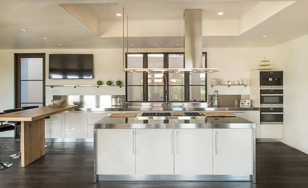 Example of a large minimalist single-wall dark wood floor eat-in kitchen design in Santa Barbara with an integrated sink, open cabinets, white cabinets, stainless steel appliances, an island, stainless steel countertops, metallic backsplash and metal backsplash