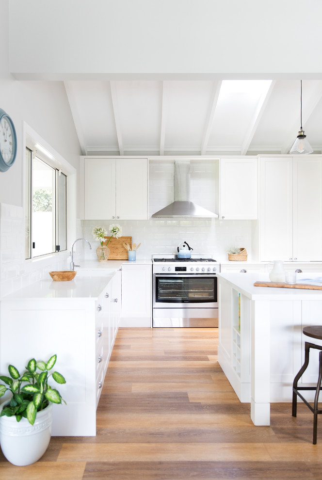 This is an example of a large farmhouse l-shaped kitchen/diner in Gold Coast - Tweed with a belfast sink, shaker cabinets, white cabinets, engineered stone countertops, white splashback, metro tiled splashback, stainless steel appliances, vinyl flooring, an island and brown floors.