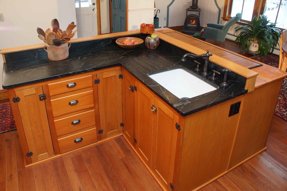 Bild på ett mellanstort amerikanskt kök, med en undermonterad diskho, luckor med infälld panel, skåp i mellenmörkt trä, bänkskiva i täljsten, stänkskydd med metallisk yta, stänkskydd i metallkakel, svarta vitvaror, mellanmörkt trägolv och en köksö