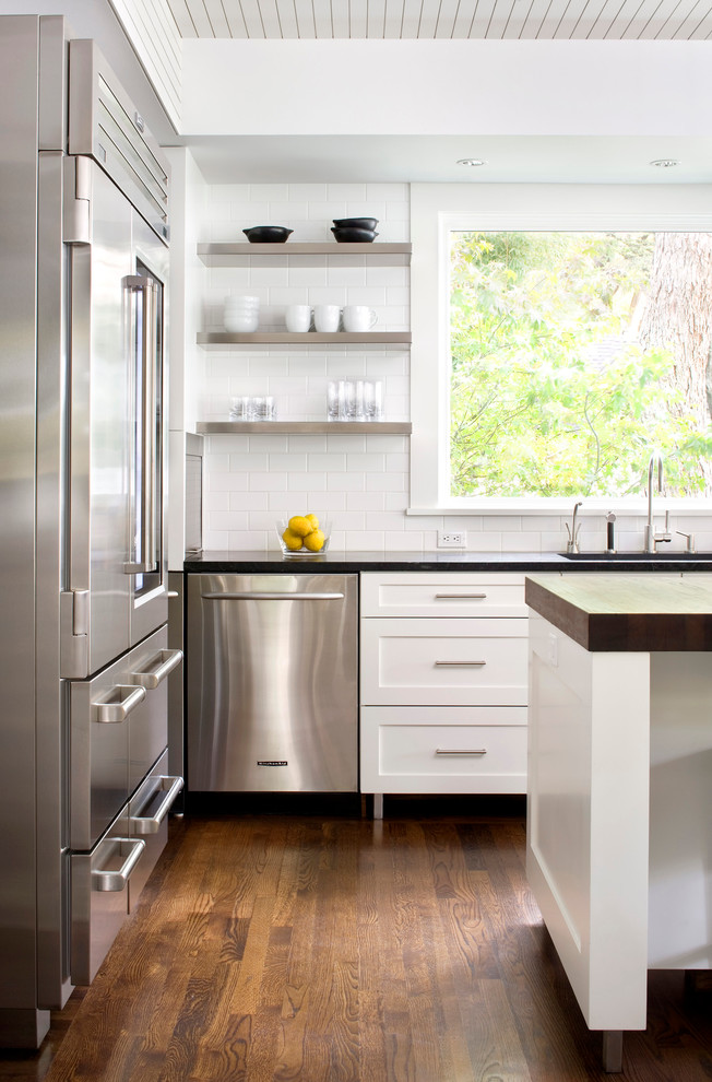 Modelo de cocina clásica renovada con armarios estilo shaker, puertas de armario blancas, salpicadero blanco, salpicadero de azulejos tipo metro y electrodomésticos de acero inoxidable